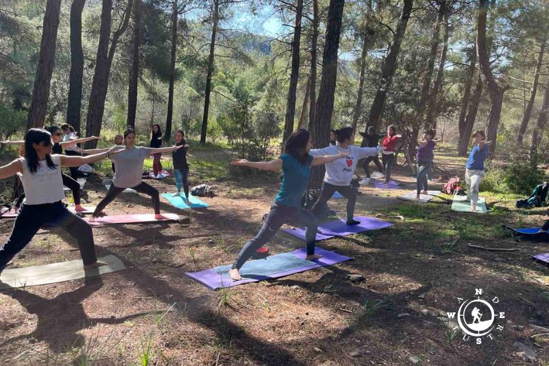 Ομάδα κάνει όρθιες στάσεις yoga με τα χέρια ανοιχτά πάνω σε στρωματάκια στο πευκοδάσος του Υμηττού