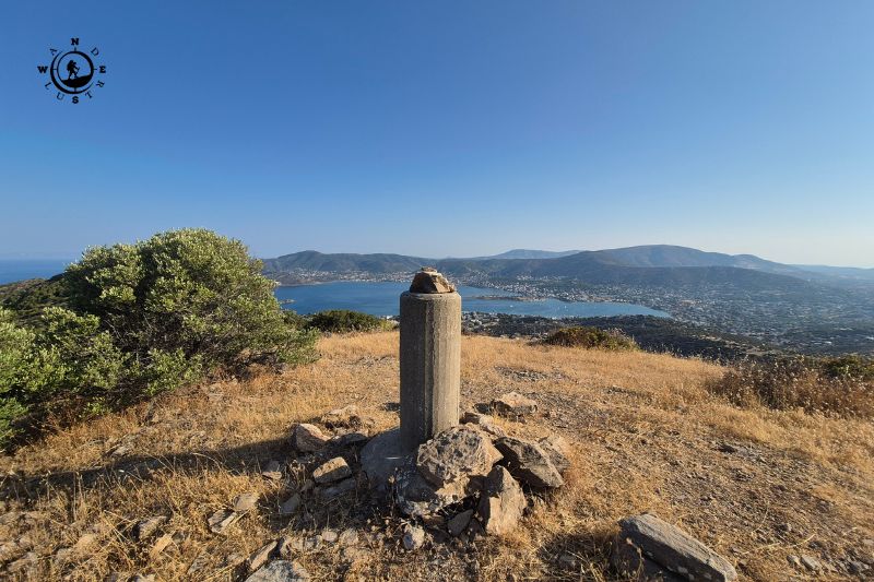 Summit marker on Perati among rocks and dry grass, with the bay in the background
