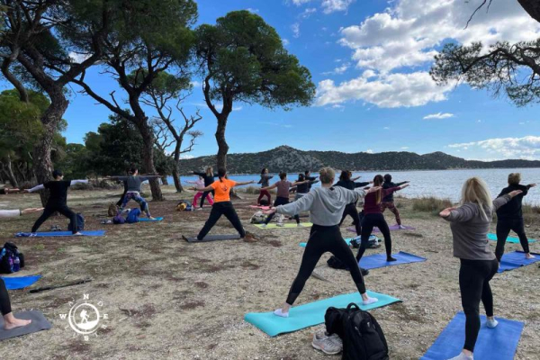 Ομάδα κάνει όρθιες στάσεις yoga πάνω σε στρωματάκια, ανάμεσα στα πεύκα και τη θάλασσα στον Σχινιά